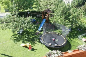 Ein Trampolin, umgeben von einer Spielanlage aus Holz mit einer Rutsche, in einer grünen Wiese mit Bäumen.