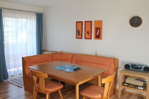 Ein Zimmer mit einem Holztisch, orangefarbenen Sitzgelegenheiten, einem kleinen Holzhochentisch mit einem Radio und gerahmten Kunstwerken an der Wand.