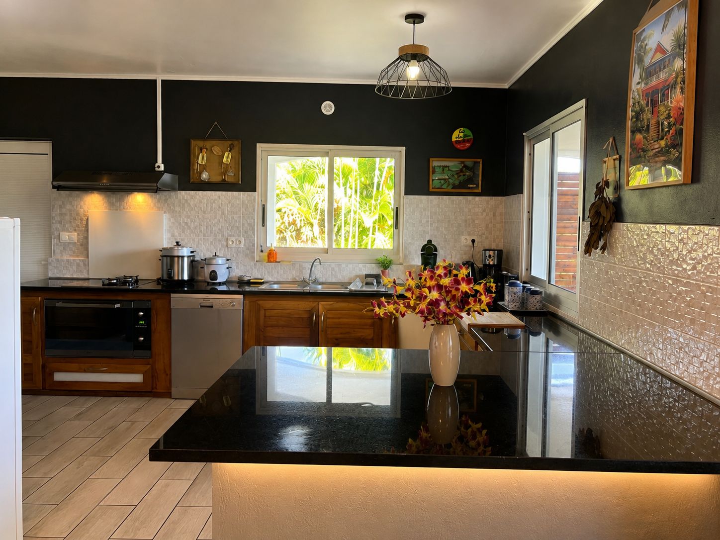 A modern kitchen with a black countertop, wooden cabinets, a window with a view of greenery, and a vase with flowers on the island.