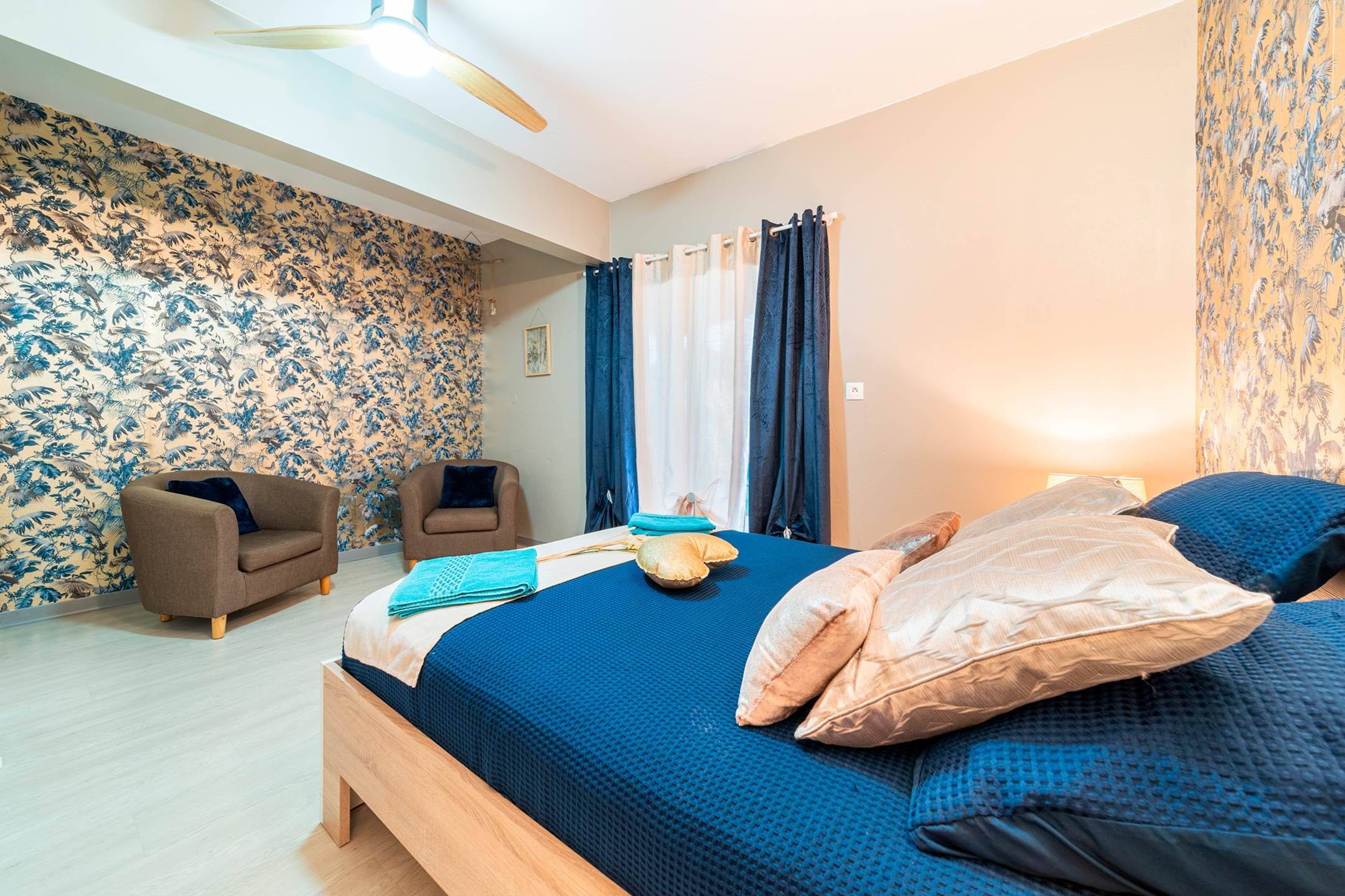 A bedroom with a bed, two armchairs, a wooden floor, and patterned wallpaper.
