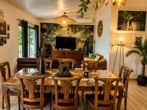 A dining area with a wooden table and chairs, a television on a wooden cabinet, and a large plant in the background.