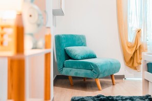 A teal armchair with wooden legs in a room with a white wall and a window with sheer curtains. - Villa Saint-Jo – Sleeps 15, ocean view, heated pool in winter (Saint-Joseph)