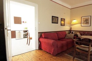 A room with a red sofa, a wooden chair, a small table, and a lamp. There is a door leading to a kitchen area with a refrigerator and a sink. - 2 Weingut Hauerhof 99 (Klosterneuburg)