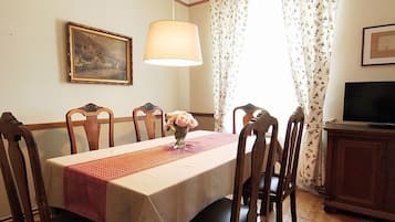A dining area with a table set for four, a vase of flowers, a painting on the wall, and a television on a cabinet.