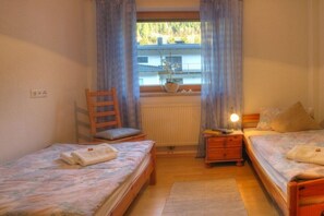 Une chambre avec deux lits simples, une chaise en bois, une table de chevet avec une lampe et une fenêtre avec des rideaux.