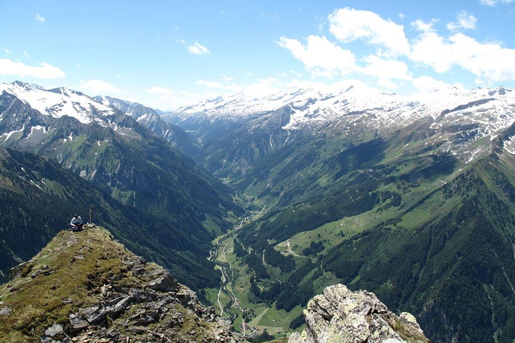 Paisagem montanhosa com um vale e uma pessoa sentada em um afloramento rochoso.