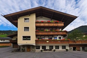 A multi-story building with balconies adorned with flowers, a stone base, and a sloped roof. - Alpenstern Fewo Tristner (Ramsau)