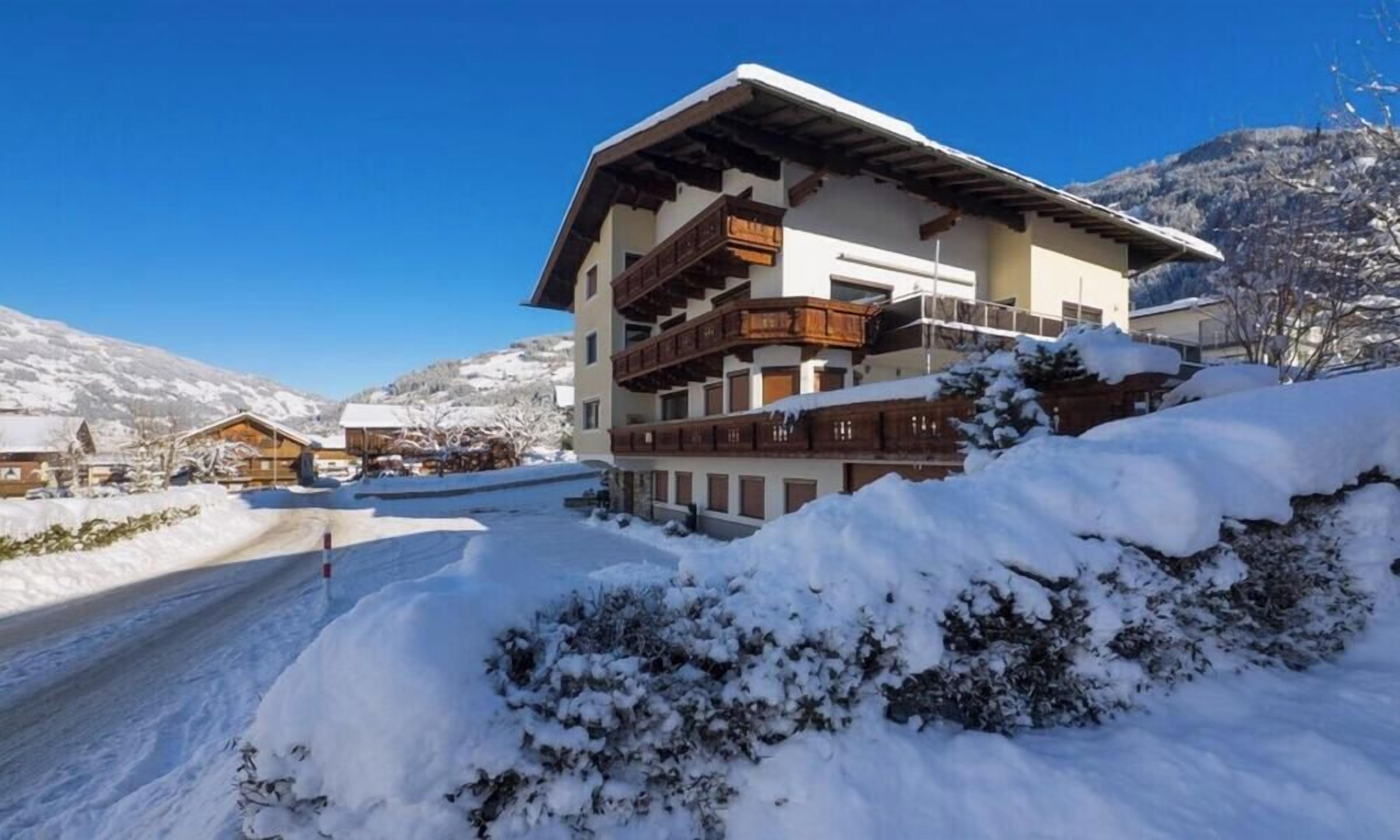A multi-story chalet with a wooden balcony, surrounded by snow-covered ground and mountains in the background.