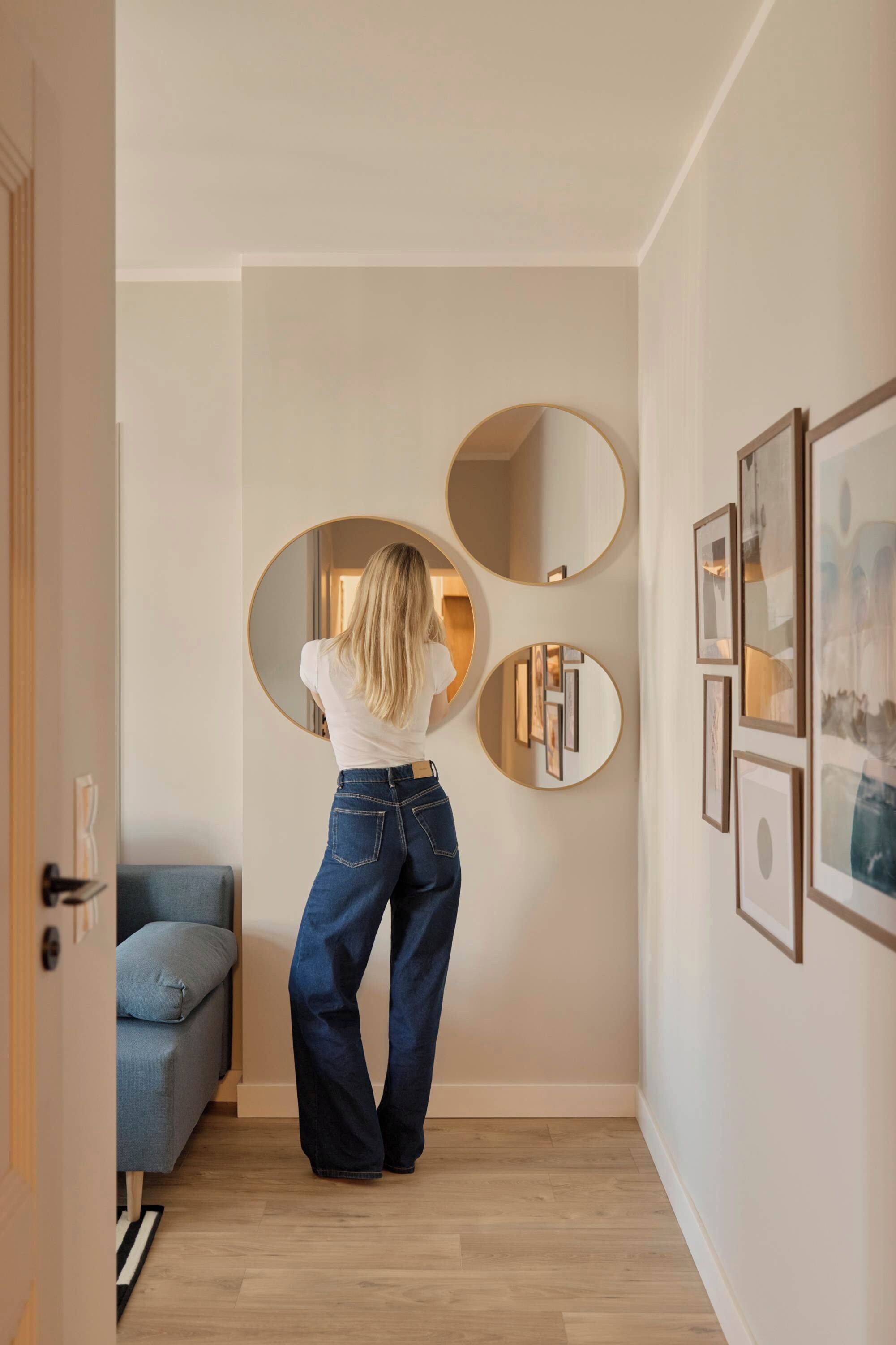 A person standing in a hallway with a door on the left and a blue sofa on the right. There are three wall-mounted mirrors and several framed pictures.