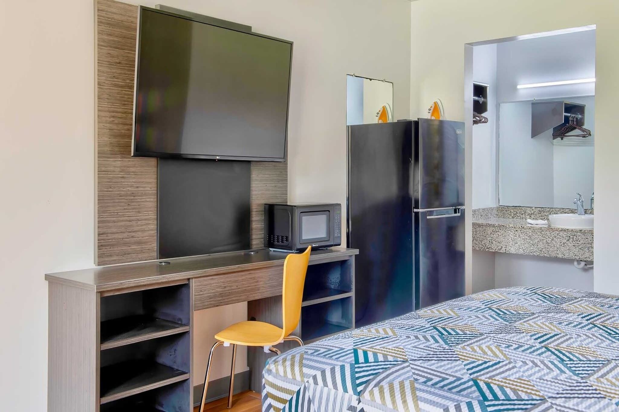 A hotel room with a bed, a desk with a chair, a television, a microwave, a refrigerator, and a bathroom with a sink and mirror.