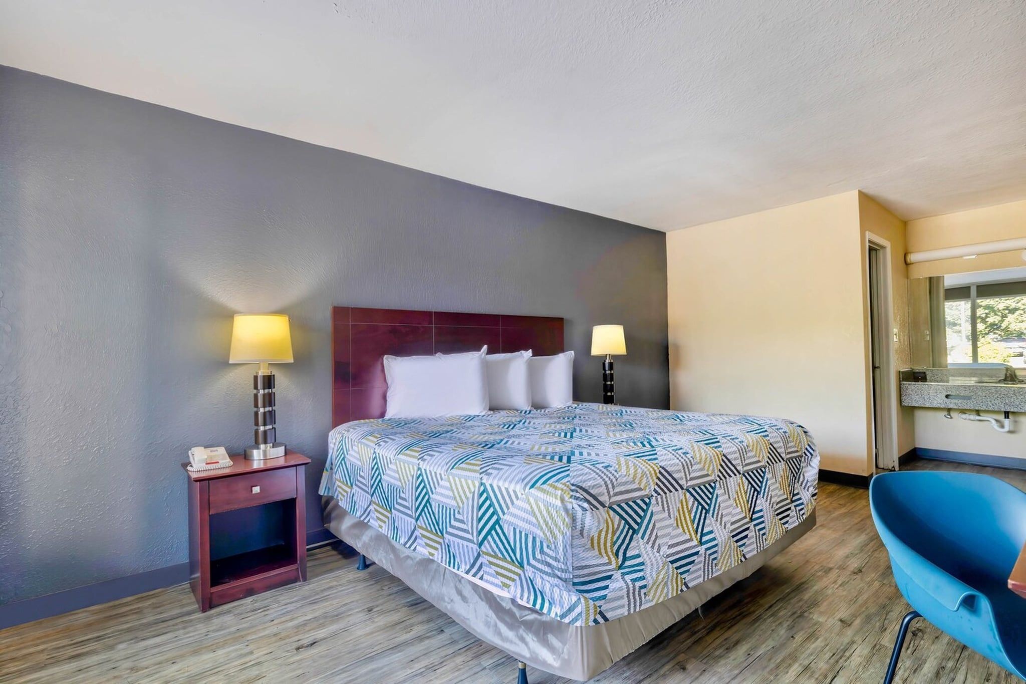 Une chambre d’hôtel avec un lit, deux lampes de chevet, une table de chevet, une chaise bleue et une fenêtre.