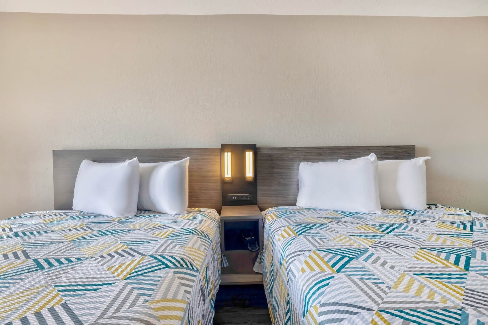 A hotel room with two beds, each with a patterned bedspread, a nightstand with a lamp, and a wall-mounted light fixture.