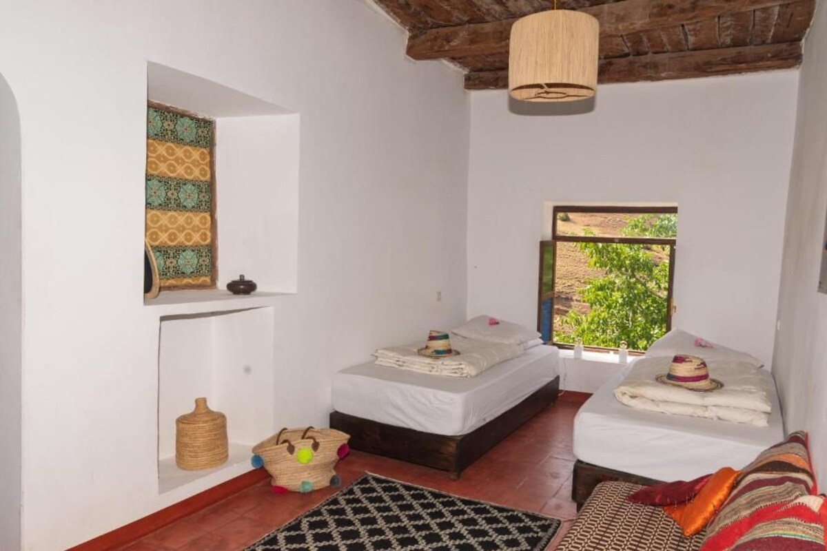 Una habitació amb dos llits, una làmpada penjant, una finestra amb vista i una cistella de veta.