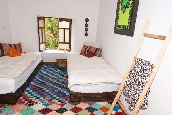 A room with two beds, a colorful rug, a window, and a wall-mounted artwork.