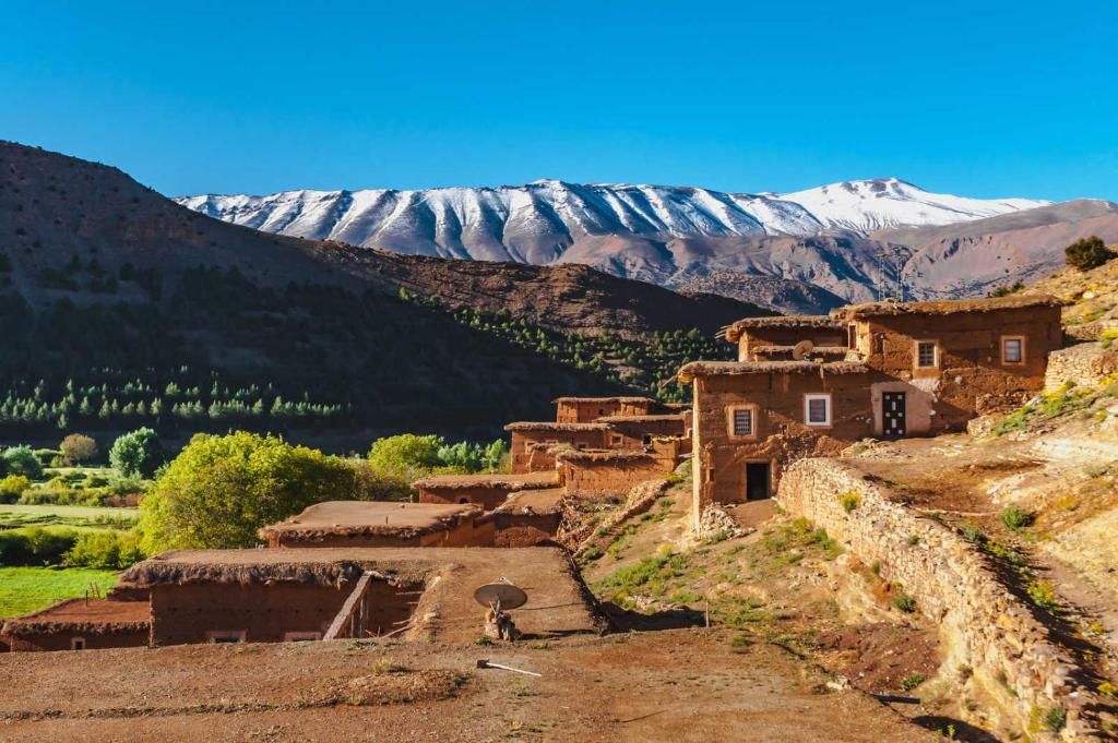 Un village traditionnel avec des maisons en adobe, un mur de pierre et une chaîne de montagnes en arrière-plan.