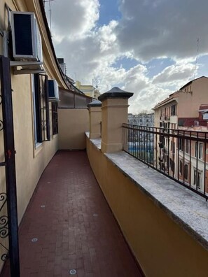 Un pasillo estrecho con suelo de baldosas, barandilla metálica y vista a edificios distantes.