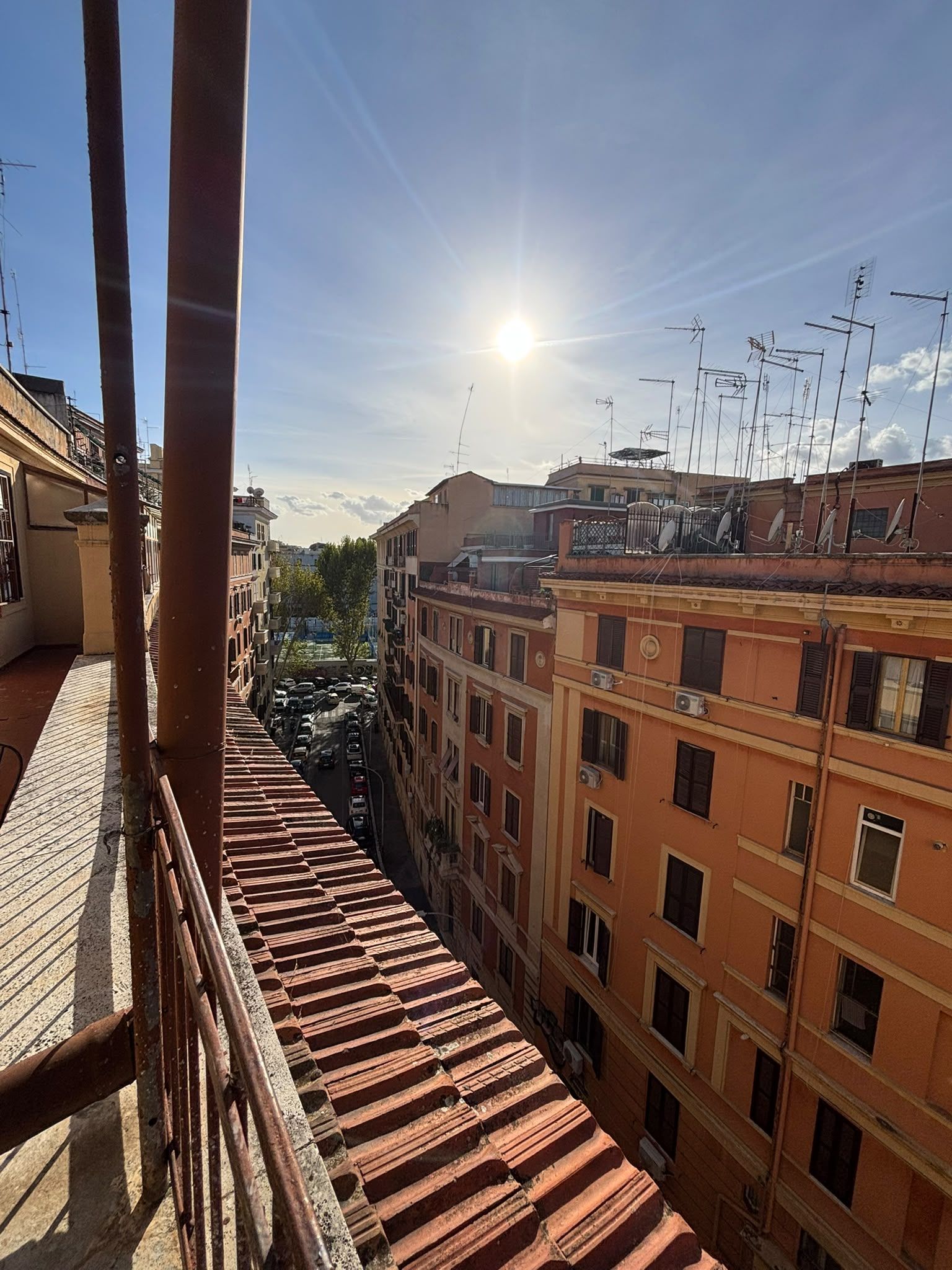 A view from a balcony overlooking a city street with buildings, parked cars, and a clear sky.
