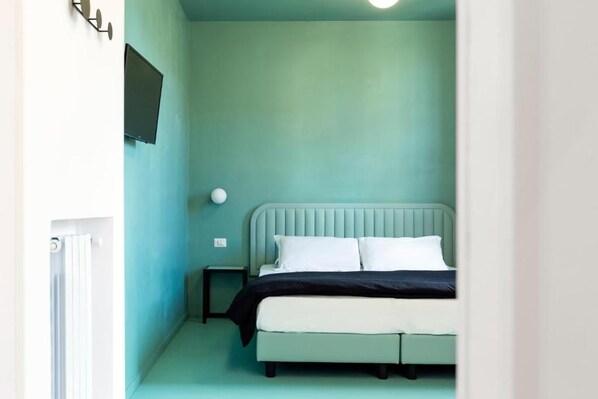 Un dormitorio con una cama, una mesita de noche, un televisor montado en la pared y un radiador.