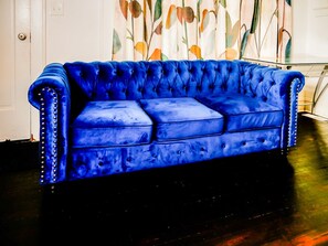 Ein blaues, mit Tupfen versehenes Sofa mit Metallnieten, platziert in einem Raum mit einer weißen Tür und einer Wand mit einem Blattmuster.