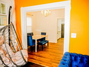 Ein Zimmer mit einem Hängekorbstuhl, einem blauen Sofa, einem Esstisch mit Stühlen und einer Chandelüster.