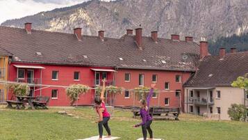 Zwei Personen üben Yoga auf Matten in einer Freiluftumgebung, im Hintergrund ein Gebäude und Berge.