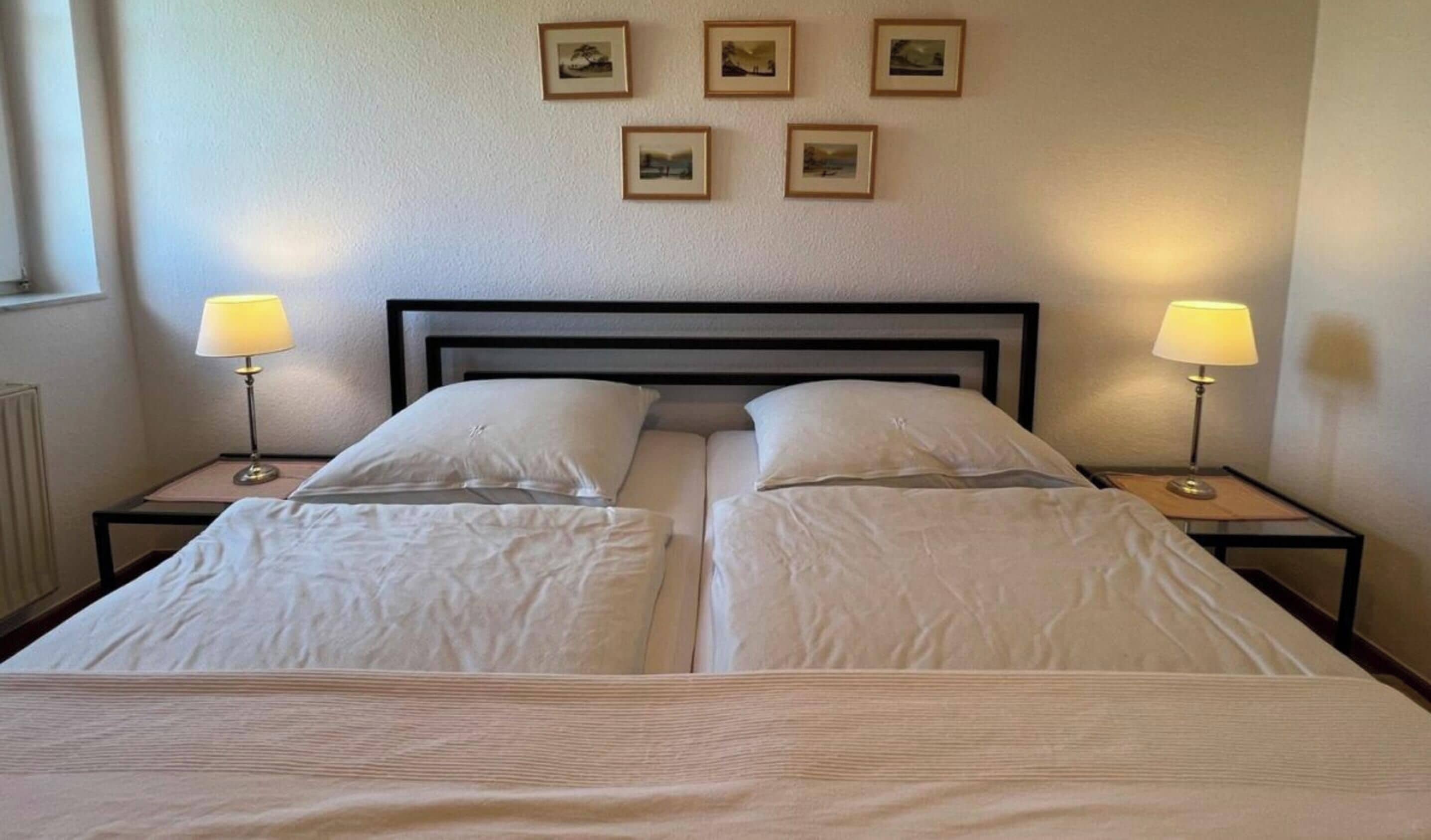 Un lit double avec deux tables de chevet, chacune équipée d’une lampe, dans une pièce dont les murs sont unis et qui est décorée de tableaux encadrés.