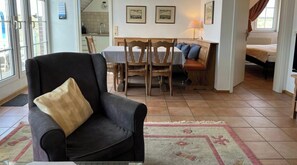 Ein Wohnzimmer mit einem dunklen Sessel, einem Esstisch mit gestreifter Tischdecke, Holzstühlen und einer durchgehende Tür erkennbaren Küche.