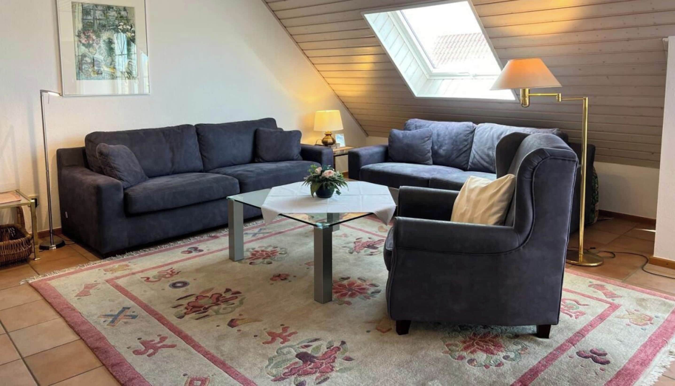 A cozy living room with a sloped ceiling, a skylight, a sofa, a chair, a small table, and a floor lamp.