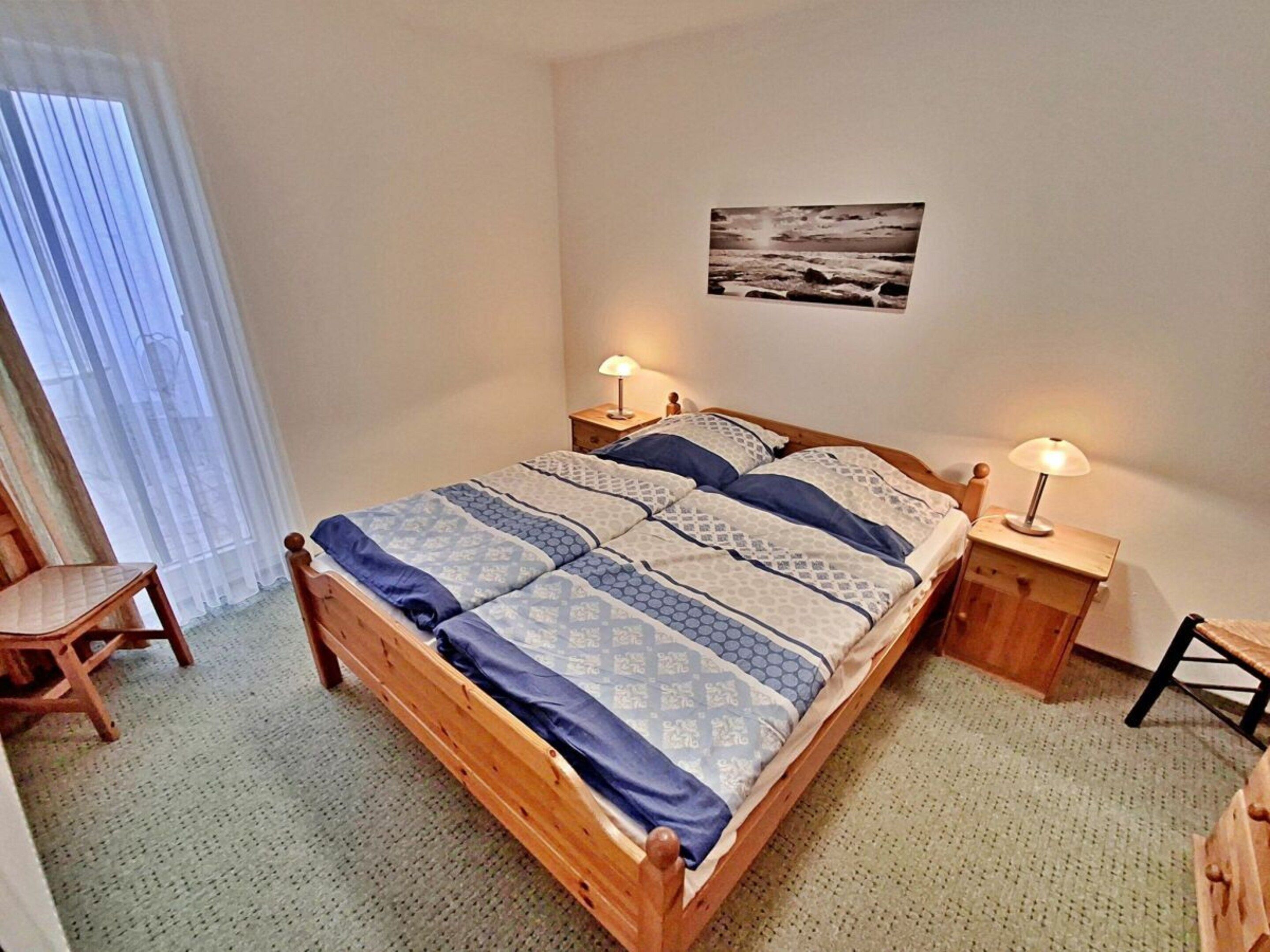 A single bed with a patterned bedspread, two bedside tables with lamps, a chair, and a wall-mounted picture.