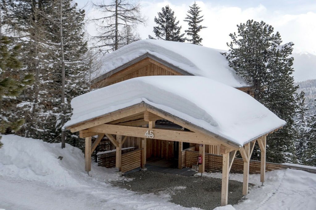 Um chalé de madeira com telhado coberto de neve, rodeado por árvores e montanhas.