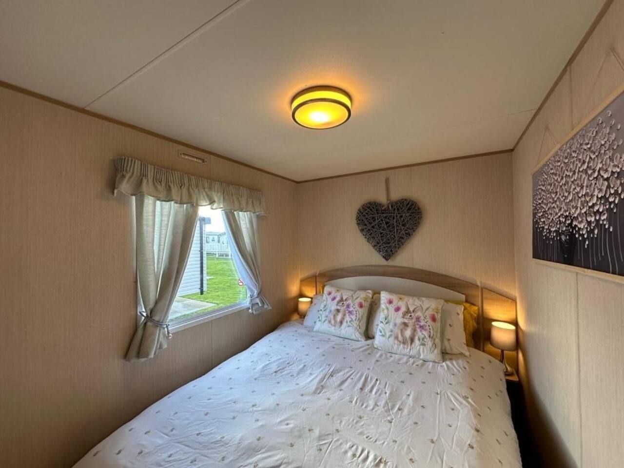 A bedroom with a bed, a window with curtains, a wall-mounted heart decoration, and a nightstand with a lamp.
