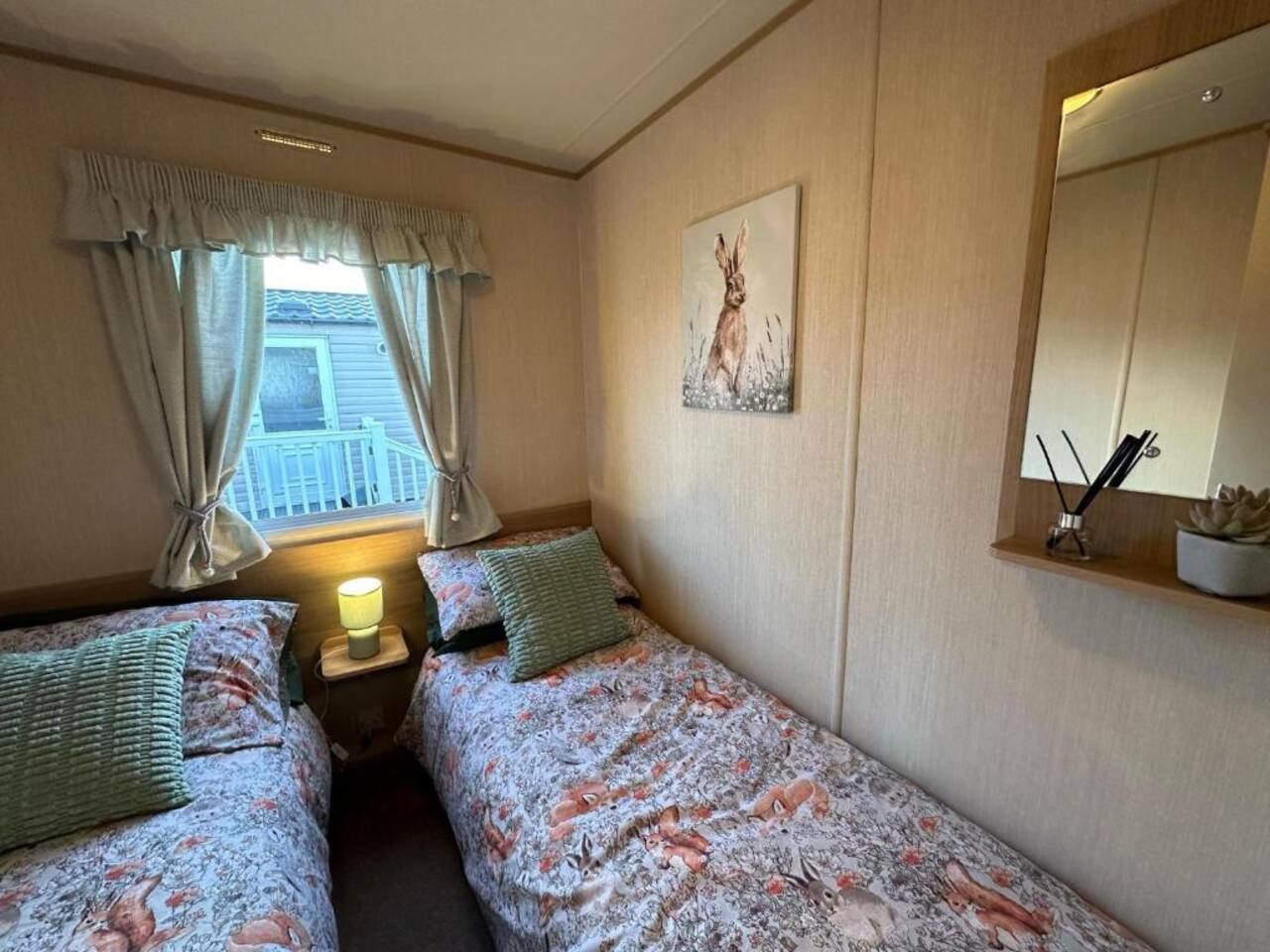 Quarto com duas camas de solteiro, janela com cortinas, um quadro na parede e mesa de cabeceira com abajur.