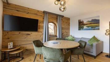 A modern living room with a wooden wall, a flat-screen TV, a round table with chairs, a grey sofa with green cushions, and a painting of a landscape.