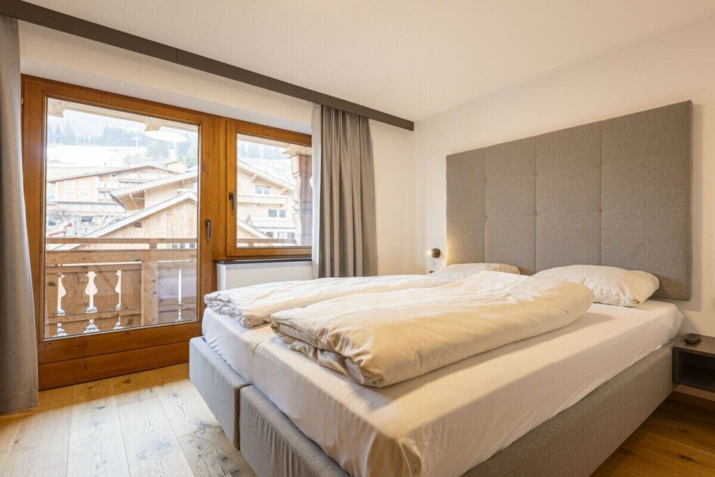 A bedroom with a double bed, a window with a view of buildings, and a wooden balcony.
