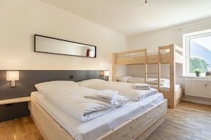 A hotel room with two beds, a bunk bed, a bedside lamp, a framed picture on the wall, and a window with a view of mountains.