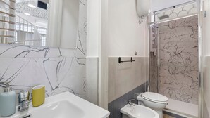Casa de banho com painel de parede em mármore, lavatório, sanita, bidé e cabine de duche com porta de vidro.