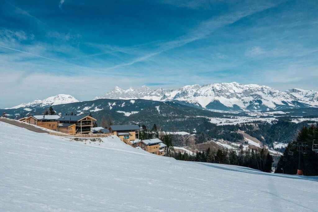 Eine verschneite Berglandschaft mit einer Berghütte, mehreren Gebäuden und einer Seilbahn im Hintergrund.