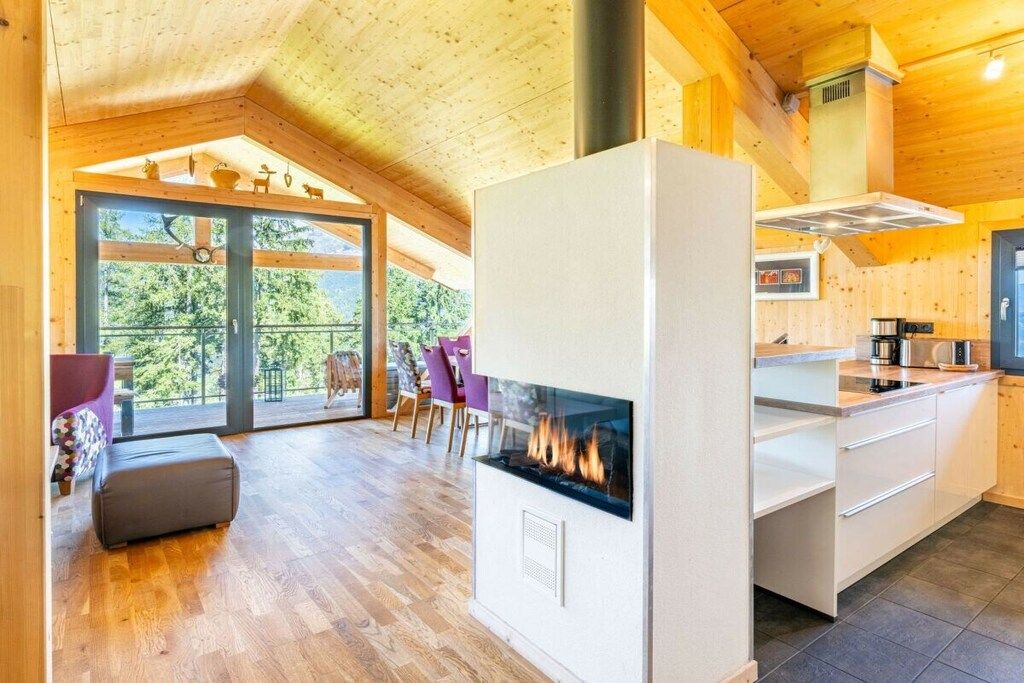 A modern kitchen with a central fireplace, wooden flooring, and a view through large glass doors.