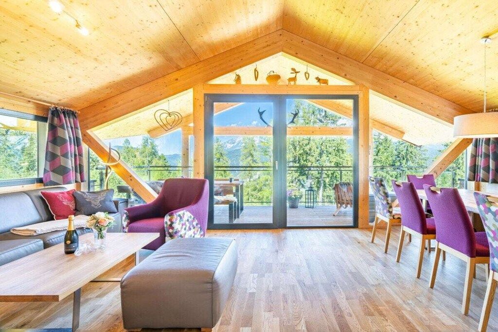 Una habitación amplia con techo alto, vigas de madera, un gran ventanal corredero que da a un espacio exterior, un sofá, una mesa con una botella y flores, y sillas moradas.