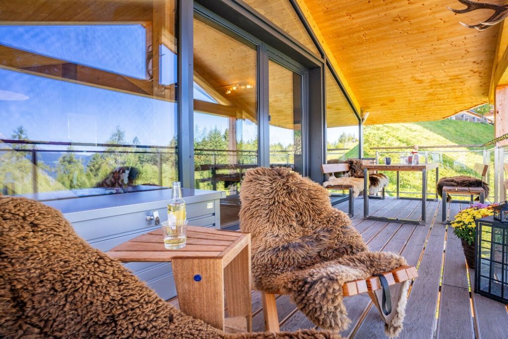 Una terraza de madera con vistas al paisaje, una mesa con una bebida en un vaso y un sillón cubierto de piel.