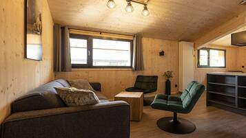 Ein gemütliches Zimmer mit einer Couch, einem grünen Sessel, einem kleinen Tisch und einem Fernseher auf einem Schrank.