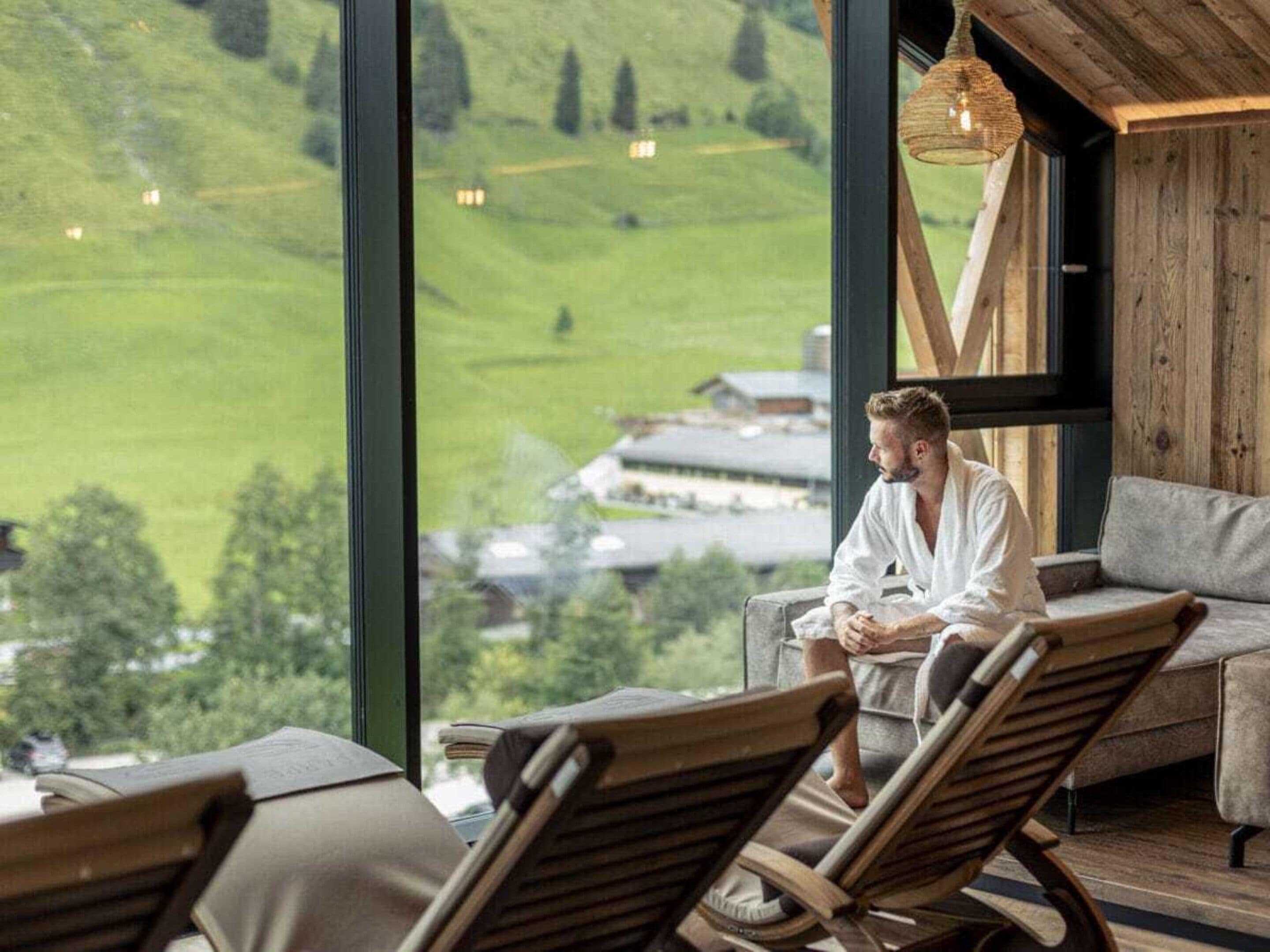 A person seated in a lounge chair with a view of a green landscape through large windows.