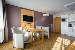 A modern room with a wooden dining table, white chairs, a flat-screen TV mounted on a wooden panel, a white ceiling light, and a kitchen area with white cabinets and appliances. - Holiday apartment # 6 with balcony (Turrach)