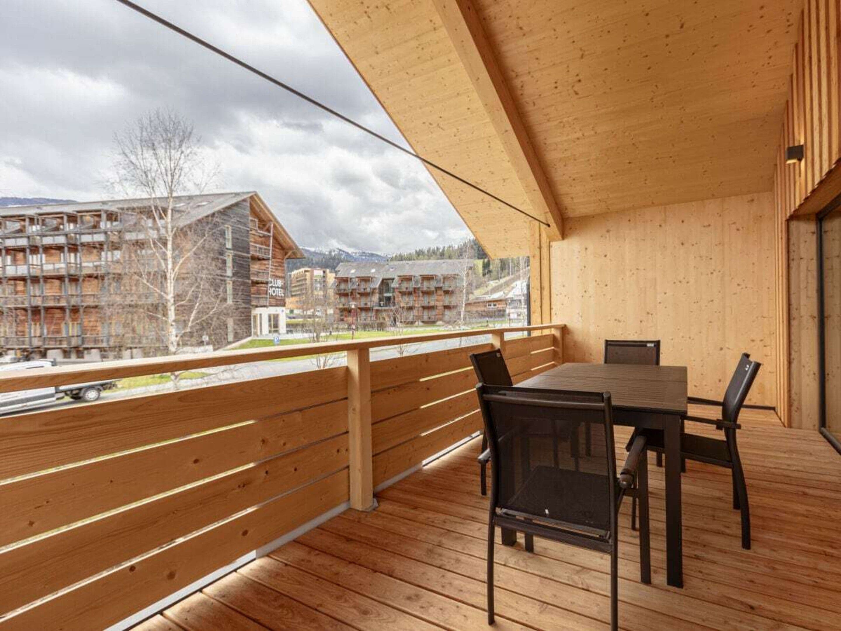 Una terraza de madera con una mesa y sillas, con vistas a una zona montañosa y edificios al fondo.