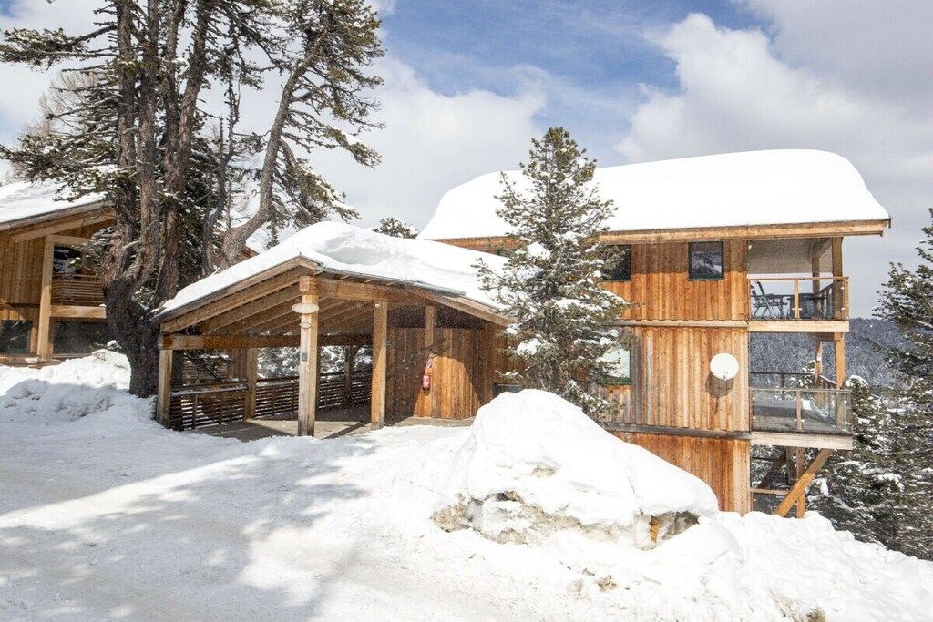 Una cabaña de madera con un porche cubierto y un balcón, rodeada de nieve y árboles.