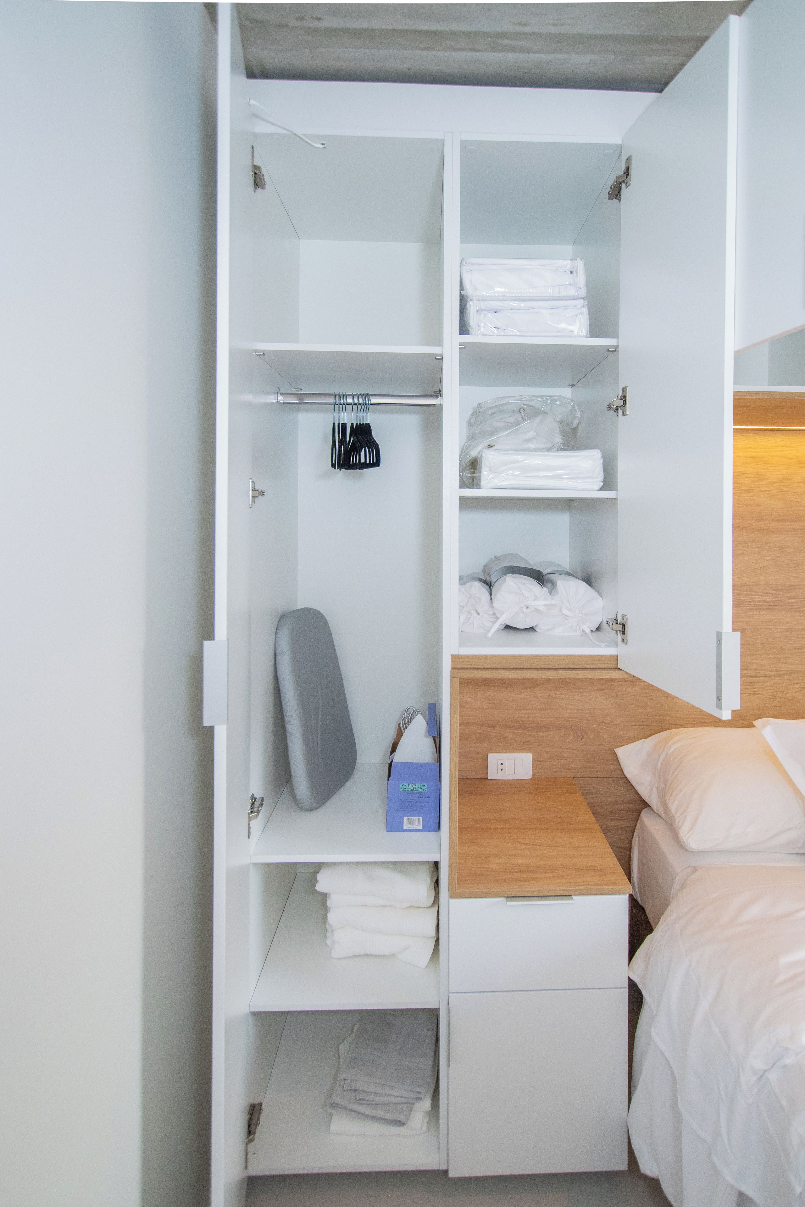A hotel room with a bed, a wooden headboard, a nightstand, a desk, and a wardrobe with shelves.