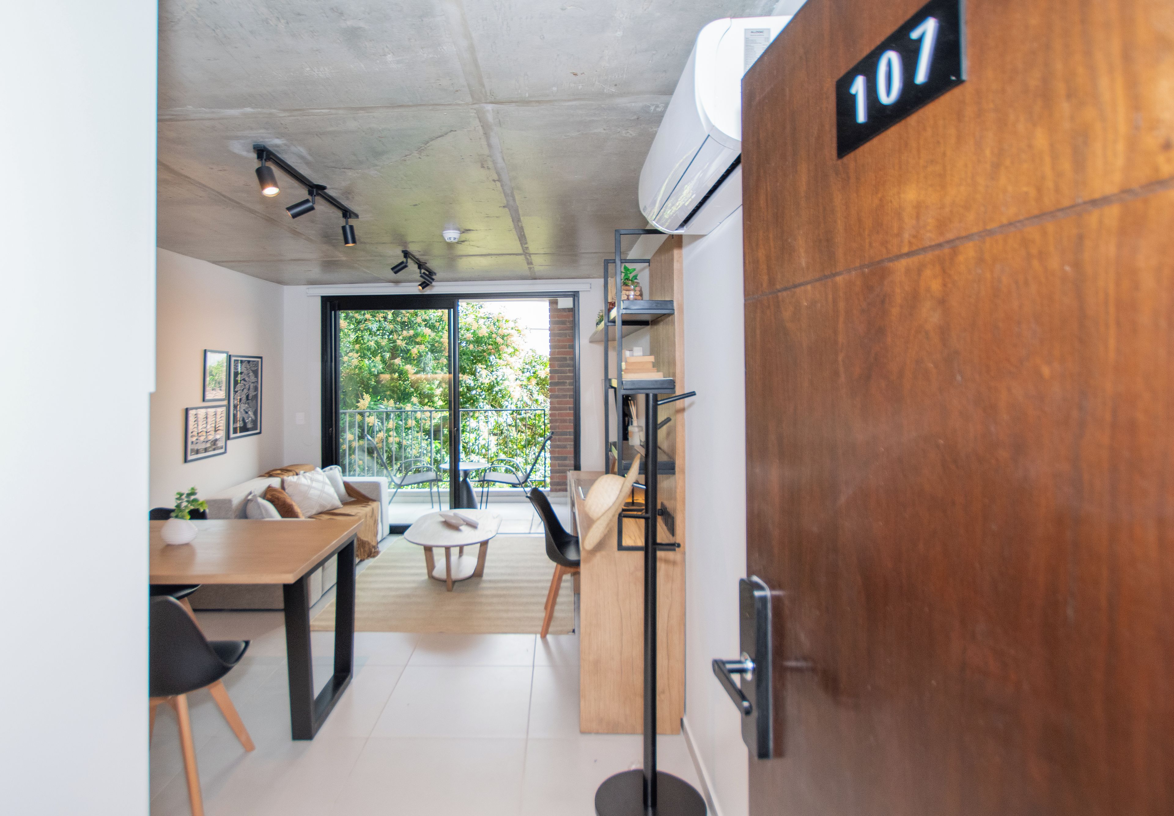 Ein modernes Interieur mit einer Holztür, auf der '107' steht, einem Essbereich mit Tisch und Stühlen sowie Blick ins Grüne durch eine Glastür.