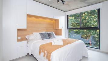 A modern bedroom with a large bed, built-in wooden headboard, and a large window offering a view of greenery.
