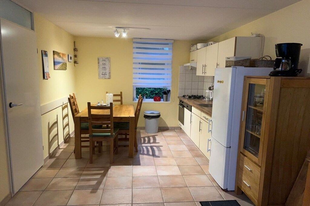A compact kitchen with a dining table, chairs, a window with blinds, a trash bin, and a refrigerator.
