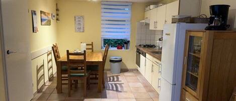 A compact kitchen with a dining table, chairs, a window with blinds, a trash bin, and a refrigerator.